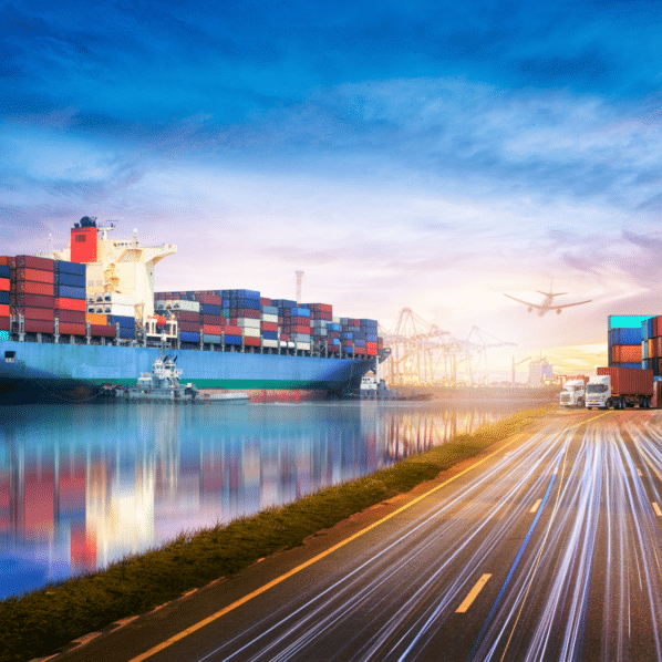 A large container ship on the water, next to a highway with semi-trucks and an airplane flying overhead