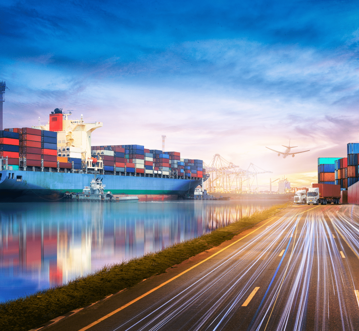 A large container ship on the water, next to a highway with semi-trucks and an airplane flying overhead